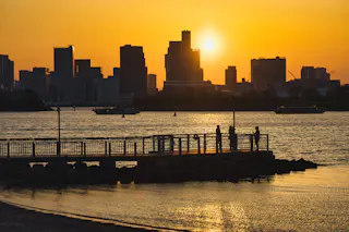 A city skyline at sunset with tall buildings silhouetted against an orange sky, a bright sun, and a pier extending into the water where people stand and look out across the river.
