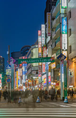 Ikebukuro A busy city street in Japan at night, filled with blurred crowds crossing the intersection. Bright neon signs in Japanese light up the buildings, creating a lively urban atmosphere.