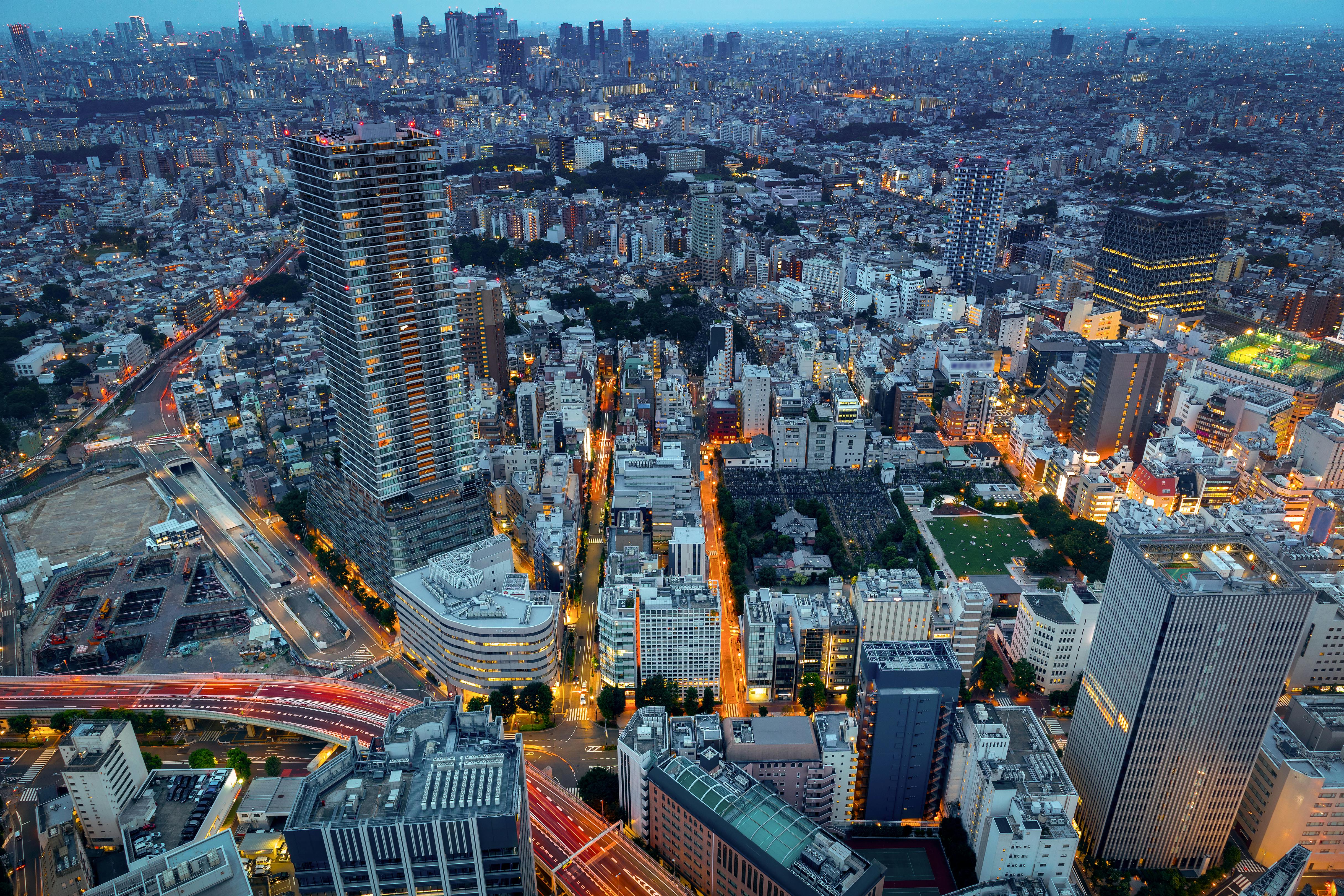 Aerial view of a dense cityscape at dusk, featuring tall buildings, illuminated streets, and a blend of modern and older architecture, with city lights beginning to glow as night approaches.