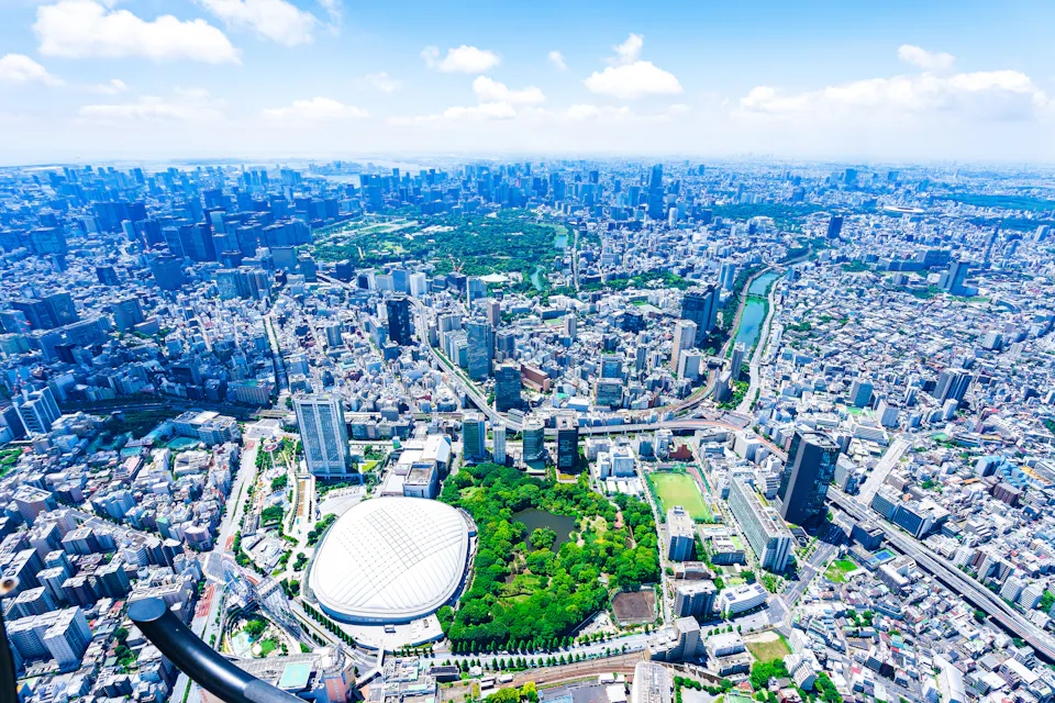 小石川後楽園の緑の木々のそばに東京ドームの白いドームが見える、東京中心部の空撮。密集した市街地が地平線まで広がっている