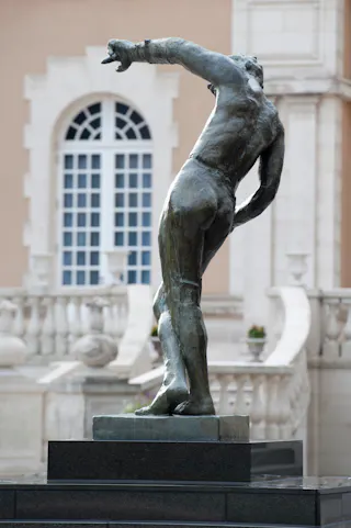 A bronze statue of a headless, muscular human figure with an outstretched arm, displayed on a pedestal in front of a classical building with arched windows and ornate stone railings.
