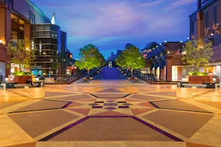 A wide, tiled plaza with an intricate geometric pattern leads to a grand staircase, flanked by modern buildings and green trees, under a colorful twilight sky.