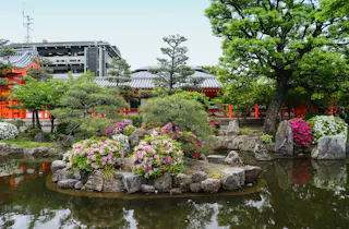A Japanese garden with a small pond, rocks, blooming pink and white azaleas, and neatly trimmed trees. A red traditional building and modern structure are visible in the background.
