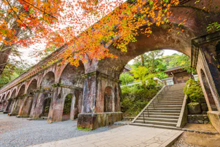 Old brick aqueduct with multiple arches under vibrant autumn foliage, next to stone steps leading upward, surrounded by trees and greenery in a tranquil, historical setting.