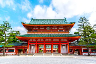A traditional Japanese temple with a green-tiled roof and vibrant red pillars, surrounded by pine trees, set against a partly cloudy blue sky.