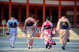 Four people wearing colorful traditional Japanese yukatas walk away from the camera towards a building with red pillars, carrying small bags.