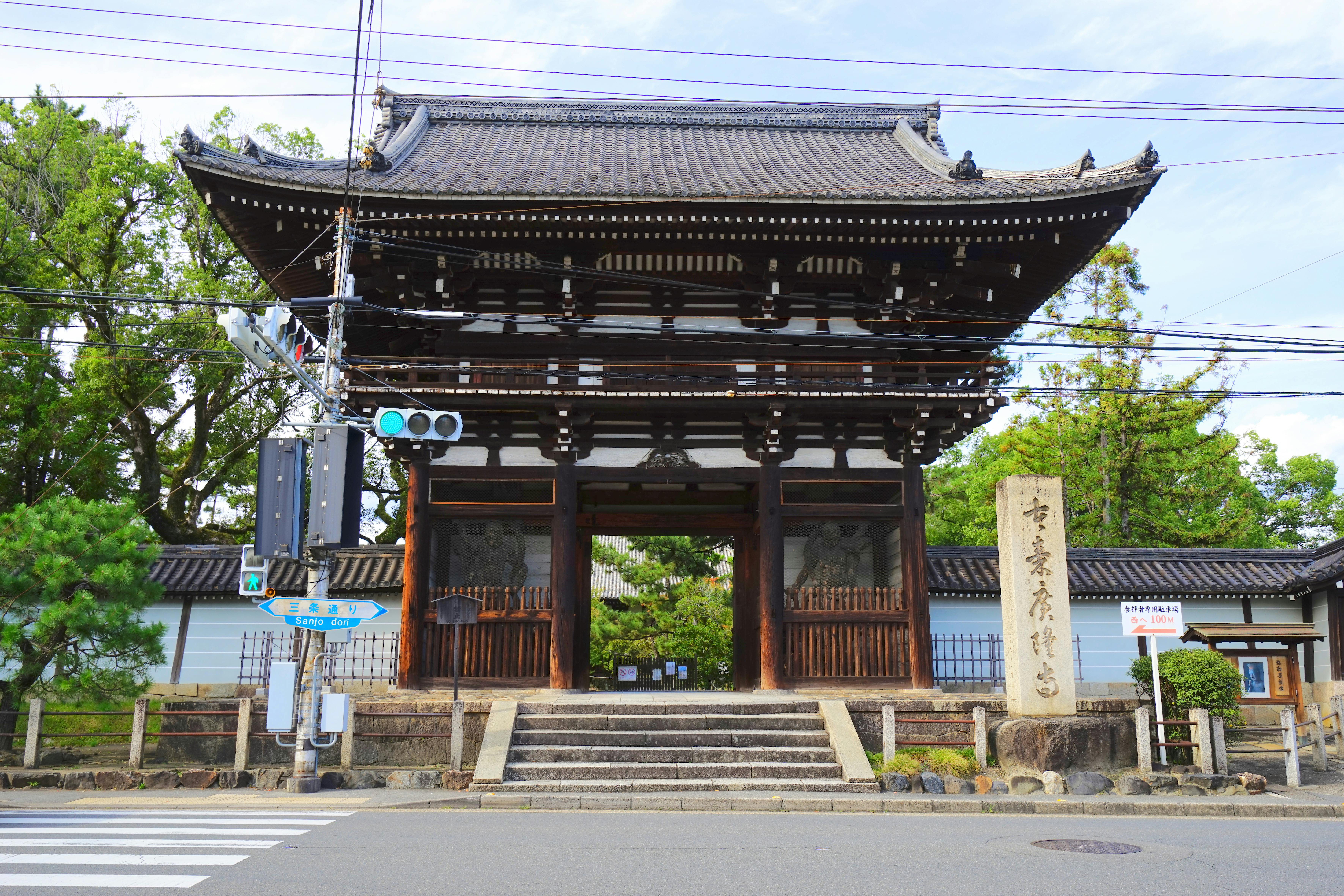 Traditional Japanese temple gate made of dark wood with a tiled roof, stone steps leading up, and statues inside. A tall stone marker with inscriptions stands beside the gate. Trees and power lines are visible in the background.