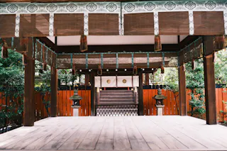 A traditional Japanese shrine with wooden flooring, hanging tassels, and ornate roof details, surrounded by greenery and an orange wooden fence. Two stone lanterns stand near the entrance to the inner sanctum.