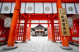 A view through bright red torii gates leading to a traditional Japanese shrine, with hanging lanterns and a wooden sign displaying Japanese characters. The courtyard and main shrine building are visible beyond the gate.