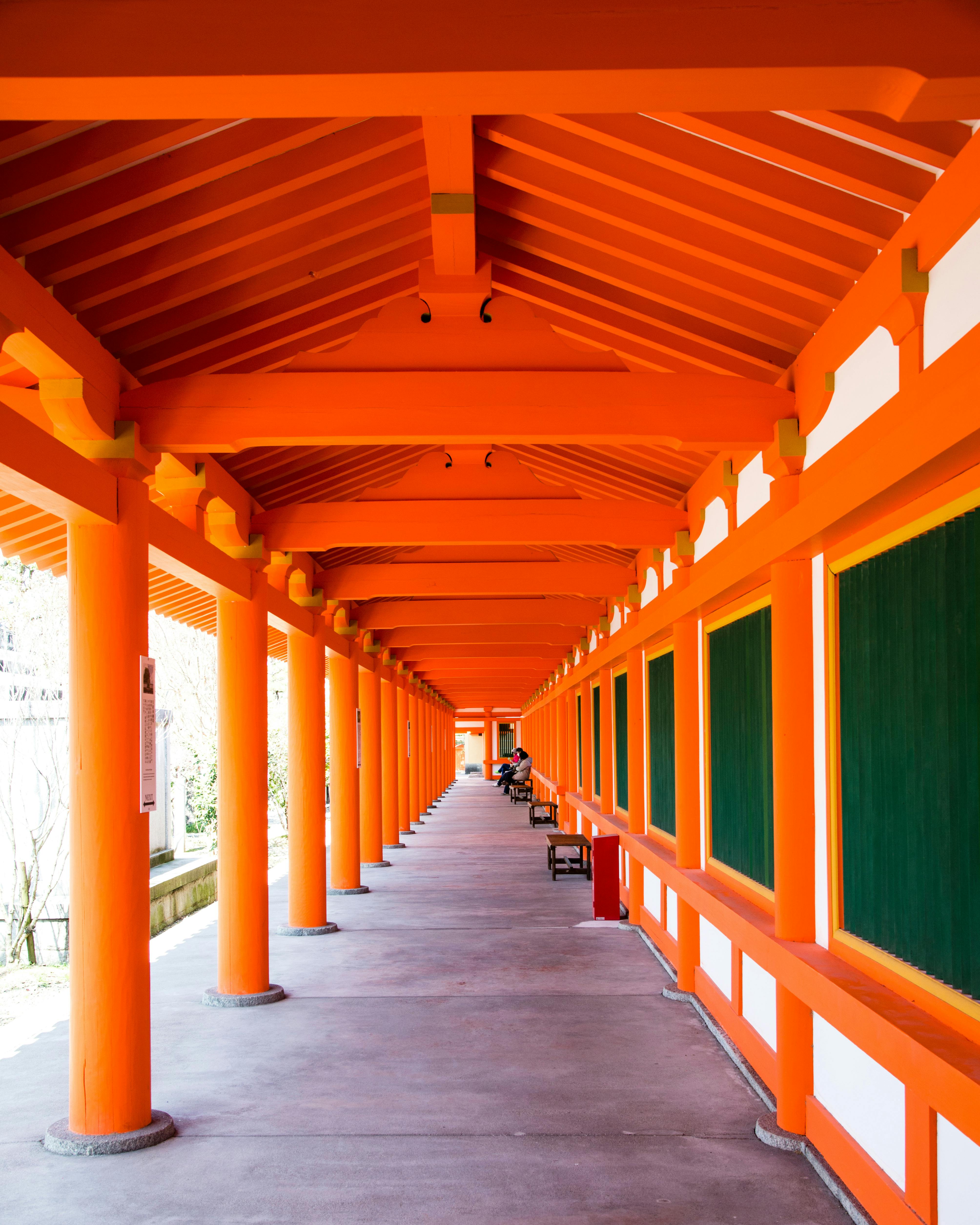 A long, covered walkway with bright orange columns and beams, green panels on the walls, and a gray floor, creates a symmetrical, open-air corridor in traditional Japanese architectural style.