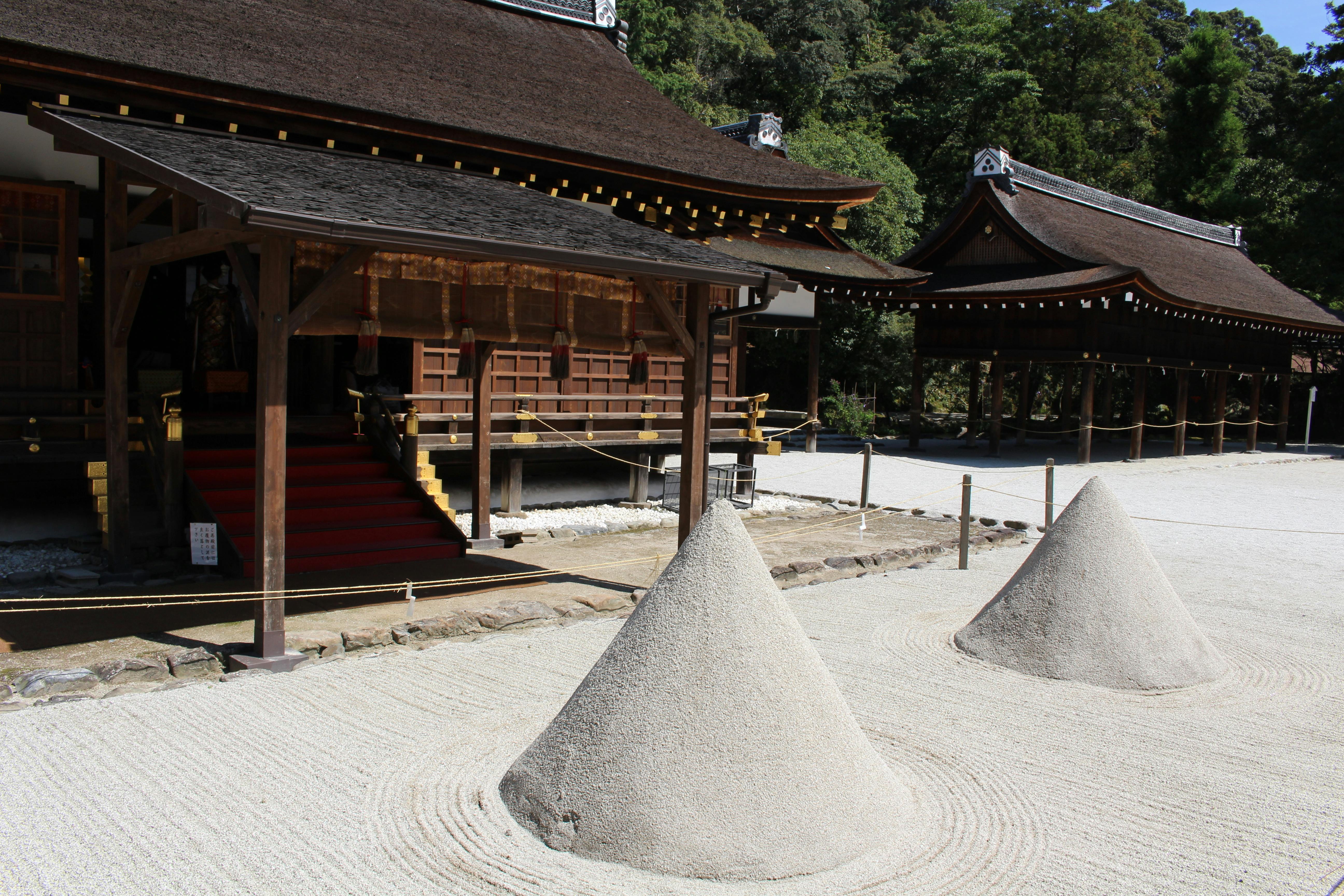 Two carefully shaped sand cones stand in a raked gravel garden outside traditional wooden Japanese buildings, surrounded by ropes and lush green trees in the background.