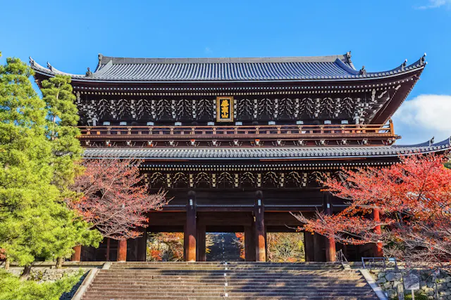 A traditional Japanese wooden temple gate stands tall, surrounded by red and green trees, with stone steps leading up to its entrance under a clear blue sky.