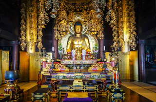 A golden Buddha statue sits at the center of an ornate altar in a richly decorated temple room, surrounded by intricate golden ornaments, colorful offerings, and traditional Japanese decor.