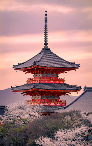 A traditional Japanese pagoda with red and white details stands amidst blooming cherry blossom trees. The sky is painted with soft hues of pink and orange, creating a serene backdrop.