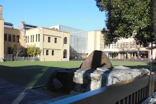 A sunlit courtyard features a large rock monument in the foreground, surrounded by modern and historic beige buildings, with a glass-walled structure in the center and a tree partially shading the right side.