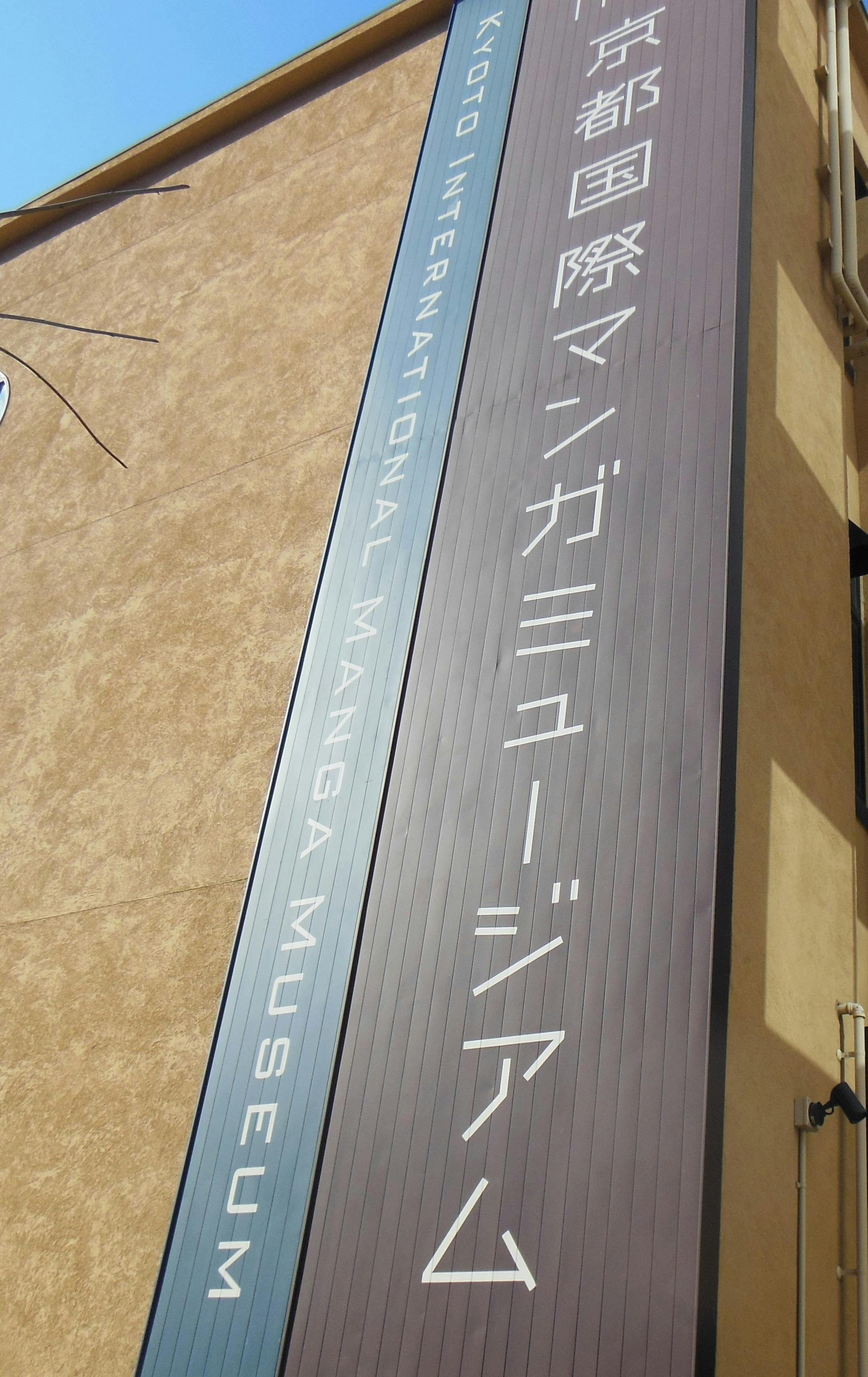 A tall vertical sign on an exterior wall reads "KYOTO INTERNATIONAL MANGA MUSEUM" in both English and Japanese. The background is beige, and the sign is dark with white lettering.