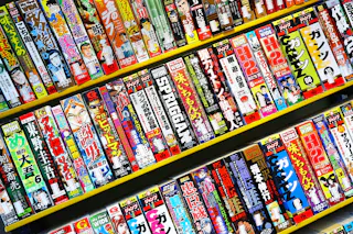 Colorful shelves filled with Japanese manga books arranged vertically, displaying vibrant covers and titles in Japanese characters, showcasing a variety of series and genres.