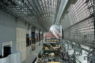A spacious modern building with a high, glass-and-steel ceiling, large open areas, escalators, and multiple levels with people walking and gathering below.