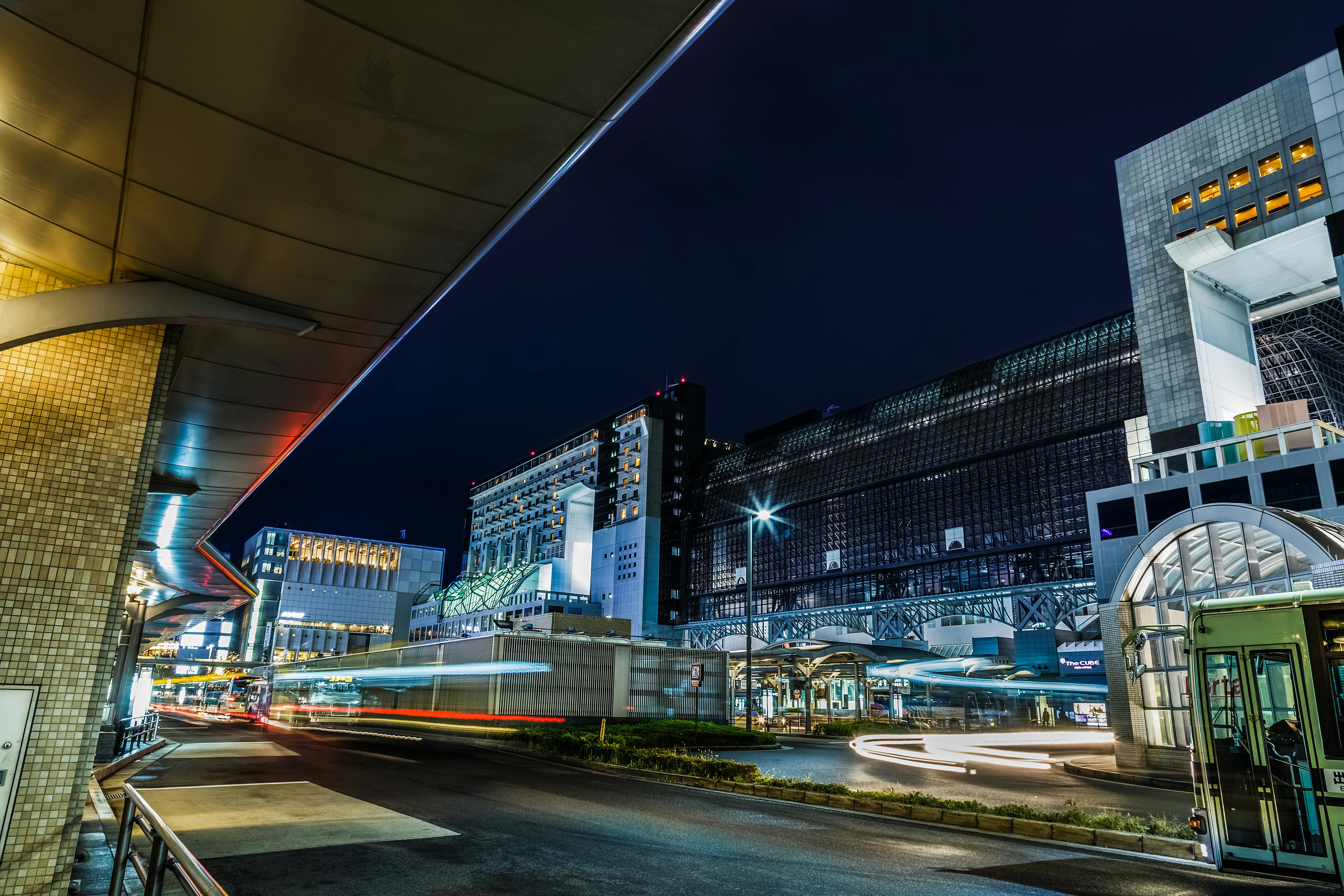 Night view of a modern cityscape with illuminated buildings, glass facades, and blurred light trails from moving vehicles on the street, creating a lively urban atmosphere.