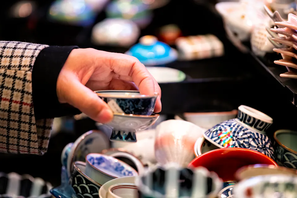 A person wearing a plaid jacket is holding a small, decorative bowl with a blue pattern among a collection of various colorful ceramic bowls on display.