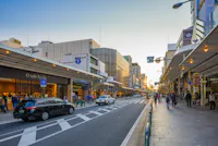 A busy city street in Japan at sunset with cars driving, people walking on sidewalks, and shops lining both sides of the road under covered walkways. The sky is clear and buildings are lit by the soft evening light.