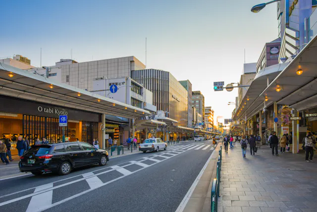 A busy city street in Japan at sunset with cars driving, people walking on sidewalks, and shops lining both sides of the road under covered walkways. The sky is clear and buildings are lit by the soft evening light.