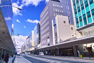 A city street lined with modern buildings and glass facades under a blue sky with clouds. Sidewalks are mostly empty, with a few vehicles and pedestrians visible.