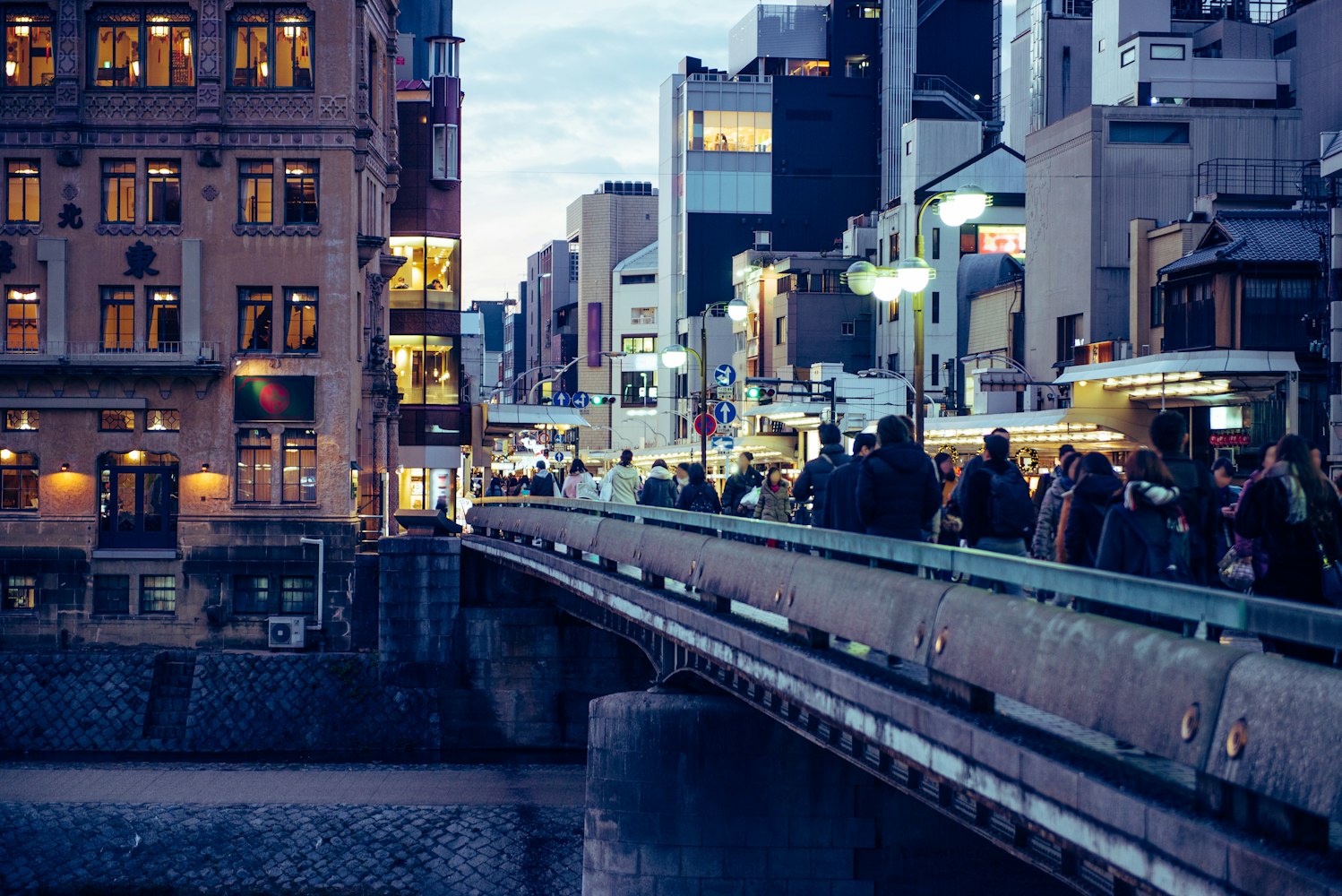 A bustling, illuminated city street at dusk with people walking along a bridge. Tall buildings line the street, adorned with various lights, creating a lively urban atmosphere. The sky is a soft blue, adding to the evening ambiance.
