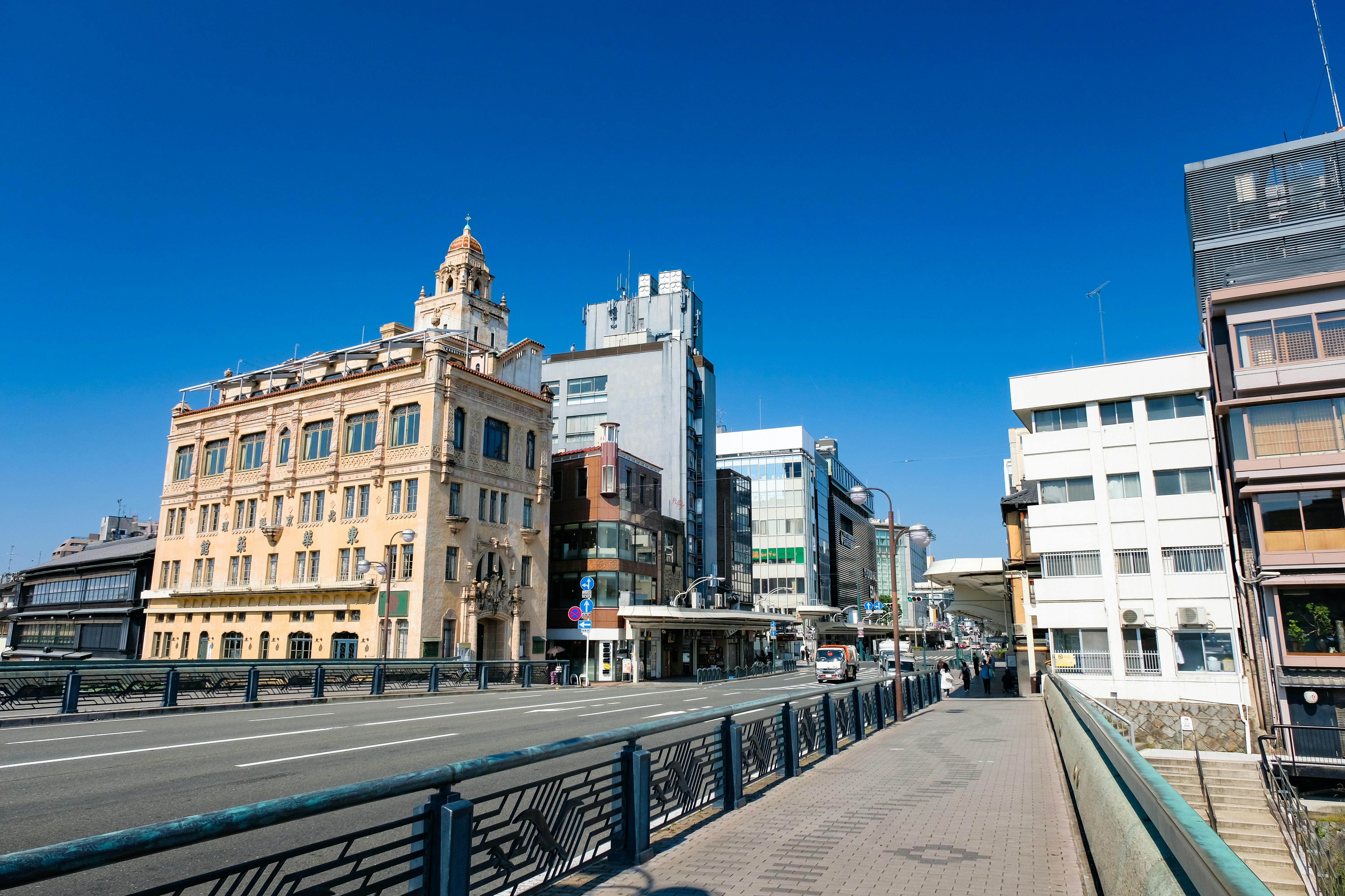 A wide street in a city with distinctive historic and modern buildings, clear blue sky, and a pedestrian walkway bordered by railings on the right side. There are few vehicles and no visible pedestrians.