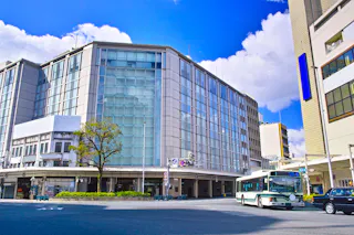 A city street corner with a modern glass building, a tree, and a green-and-white city bus stopped at a bus stop under a bright blue sky with clouds.