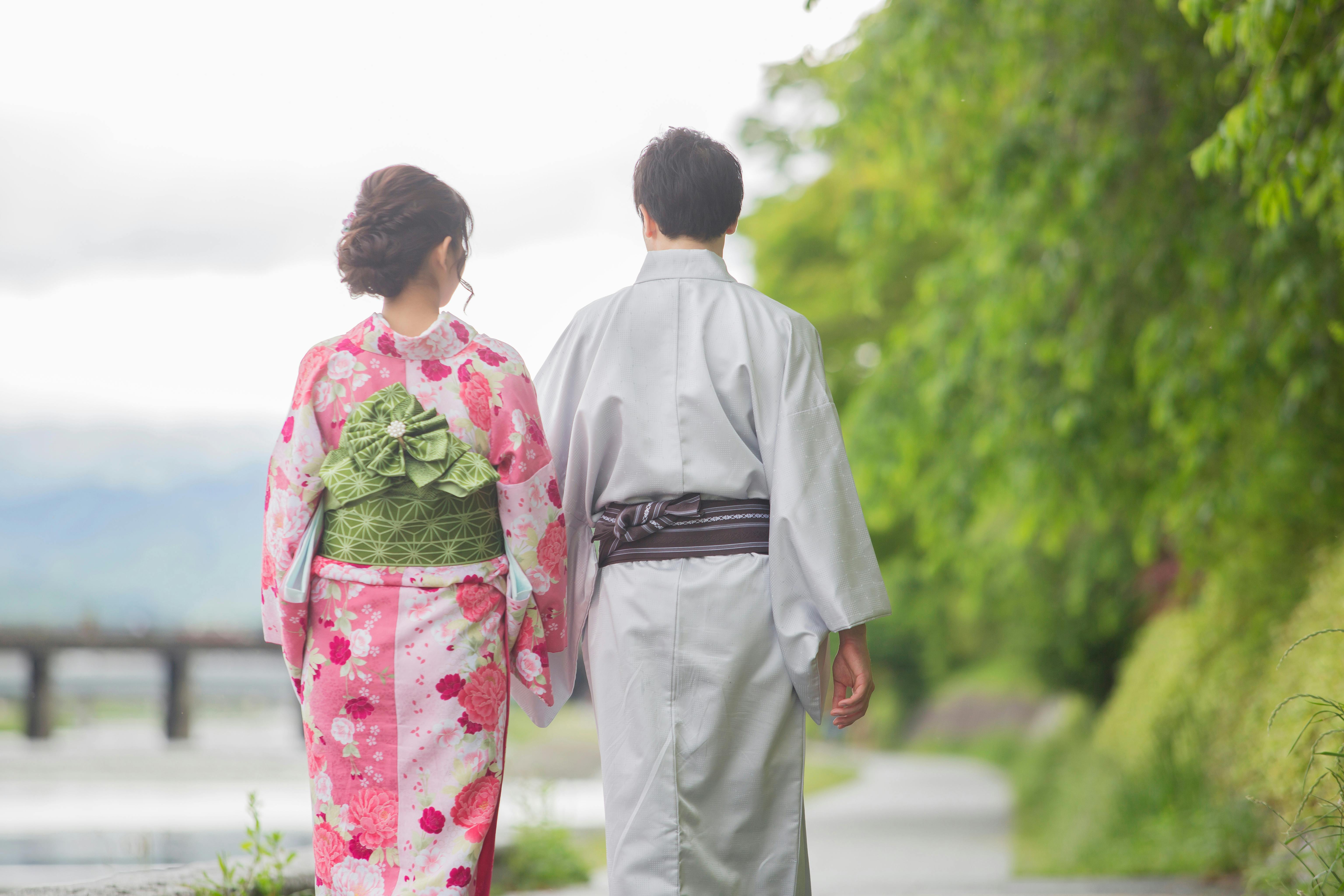 A woman and a man wearing traditional Japanese clothing walk side by side on a path near greenery and water, seen from behind.