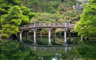A stone arch bridge with decorative railings spans a calm pond, surrounded by lush green trees and reflected clearly in the water below.