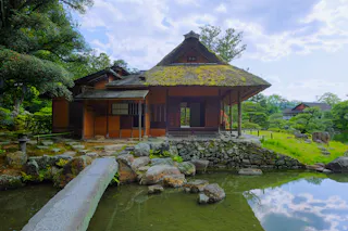 A traditional Japanese tea house with a thatched roof stands beside a pond, surrounded by lush greenery and stone pathways under a partly cloudy sky.