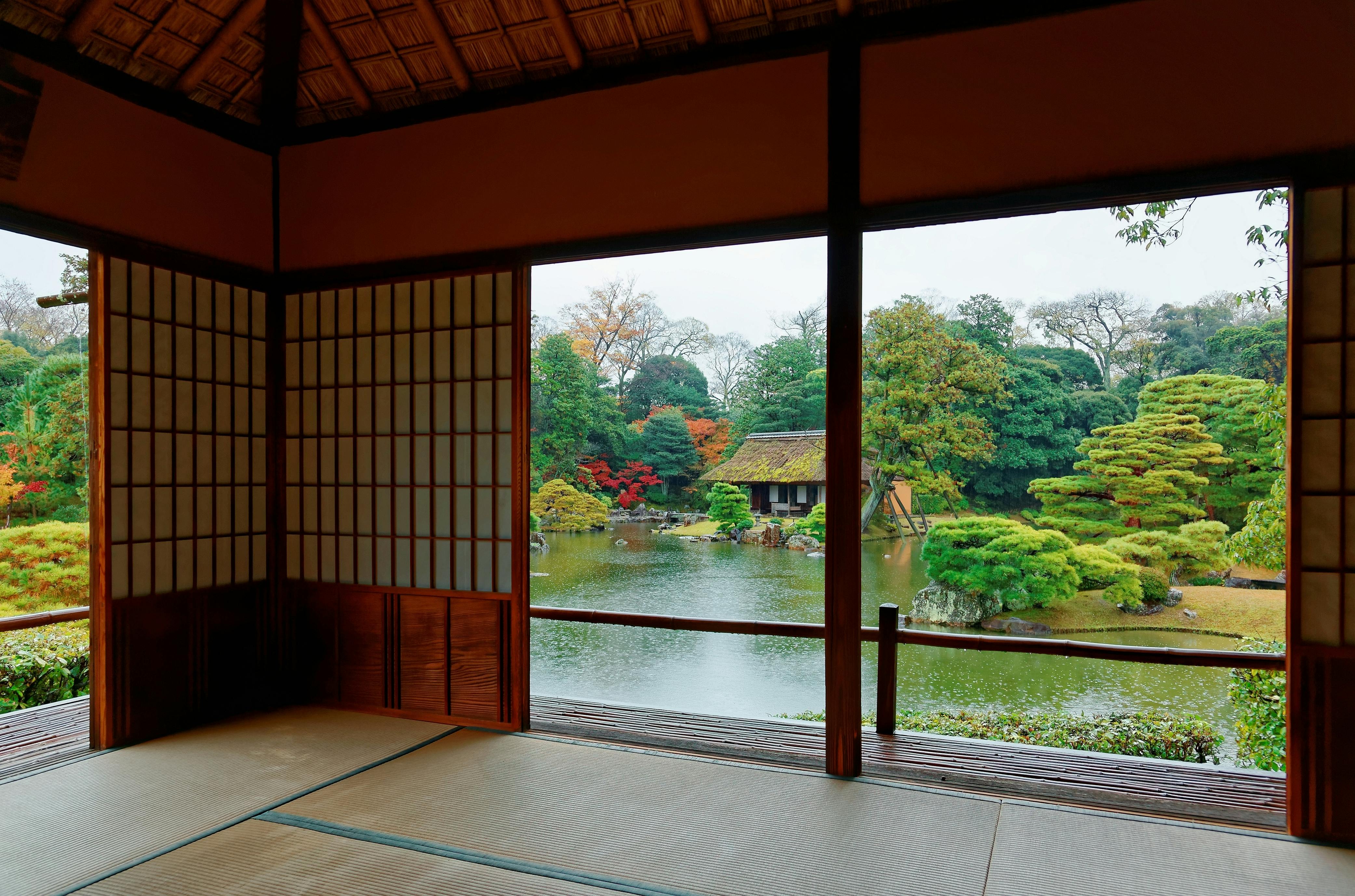 View from inside a traditional Japanese room with tatami mats and shoji screens, looking out onto a serene garden with a pond, lush greenery, and a small wooden tea house.