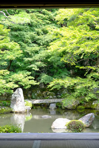 A serene Japanese garden with lush green trees, large rocks, and a tranquil pond reflecting the greenery, viewed from under a shaded structure.