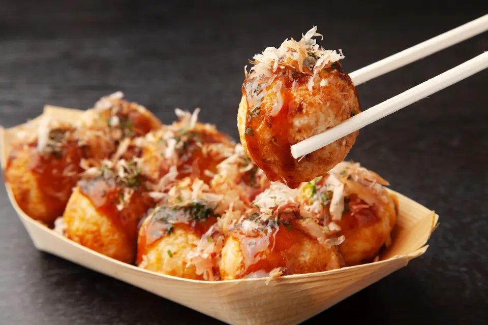 A pair of chopsticks holding a piece of takoyaki topped with sauce and bonito flakes over a wooden tray filled with more takoyaki balls. The background is a dark surface.