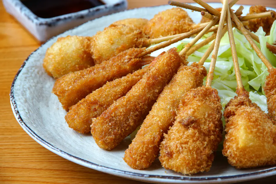 A plate of assorted kushiage, which are deep-fried skewers coated in breadcrumbs, arranged in a fan shape. They're accompanied by shredded cabbage and a small dish of dipping sauce in the background.