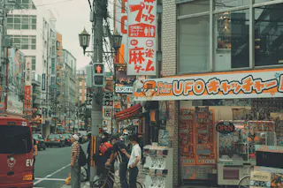 A busy street scene in Japan with people waiting at a crosswalk, surrounded by colorful shop signs and buildings, including one advertising a UFO catcher arcade. A red bus is visible on the left.