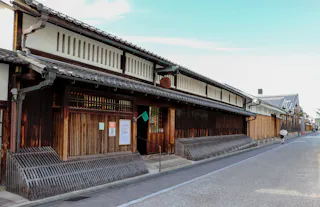 A traditional Japanese wooden building with tiled roof lines a quiet street under a clear sky; a person with an umbrella walks in the distance.