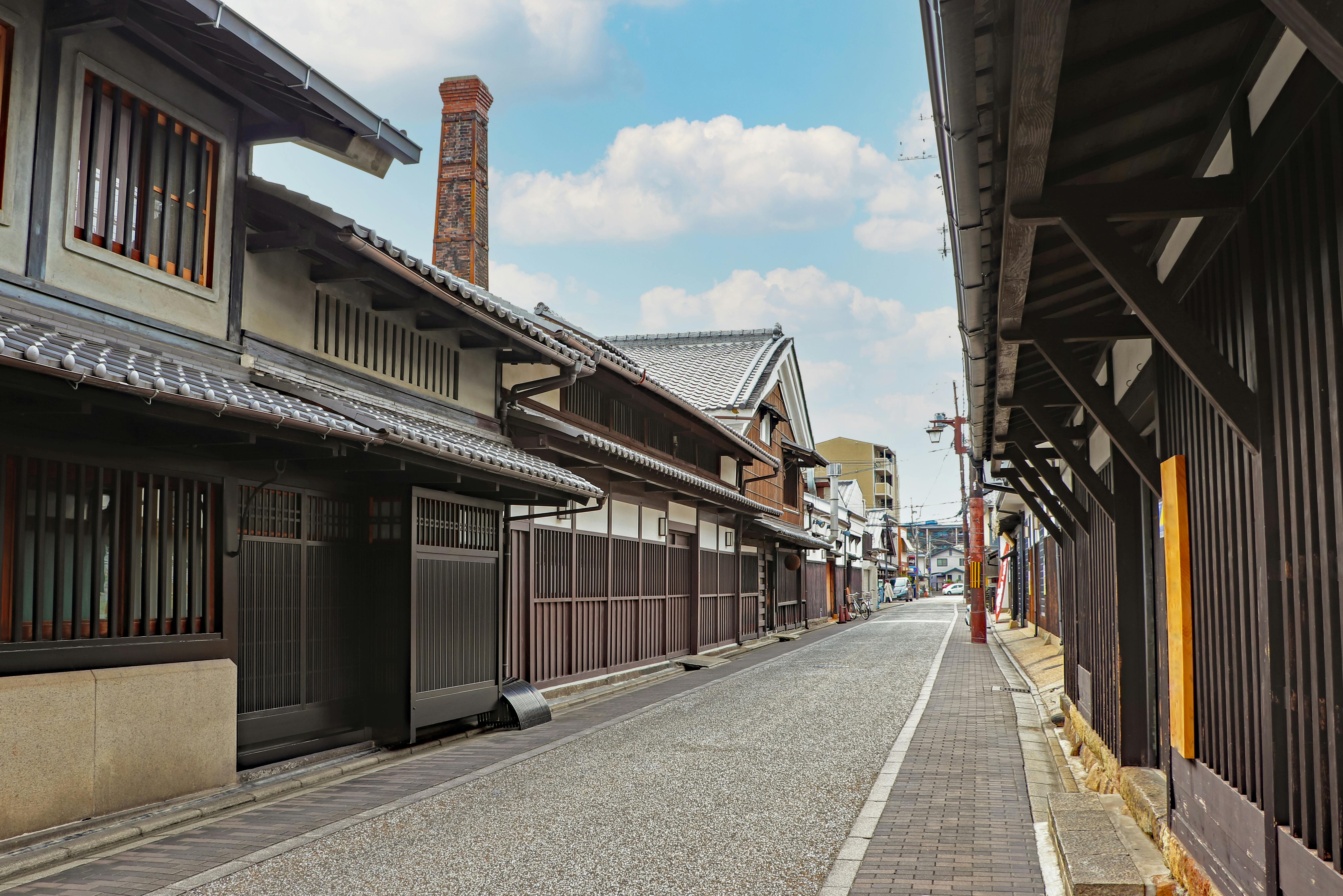 A quiet street lined with traditional Japanese wooden buildings, featuring tiled roofs and wooden lattice windows, under a partly cloudy sky. No people or vehicles are visible.
