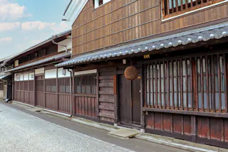 A traditional Japanese street with wooden buildings, sliding doors, and barred windows, featuring a brown sugidama (cedar ball) hanging above a doorway under a tiled roof.