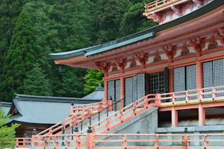 A traditional Japanese temple with orange and white wooden architecture, elevated stairs, and intricate details, surrounded by lush green trees in the background.