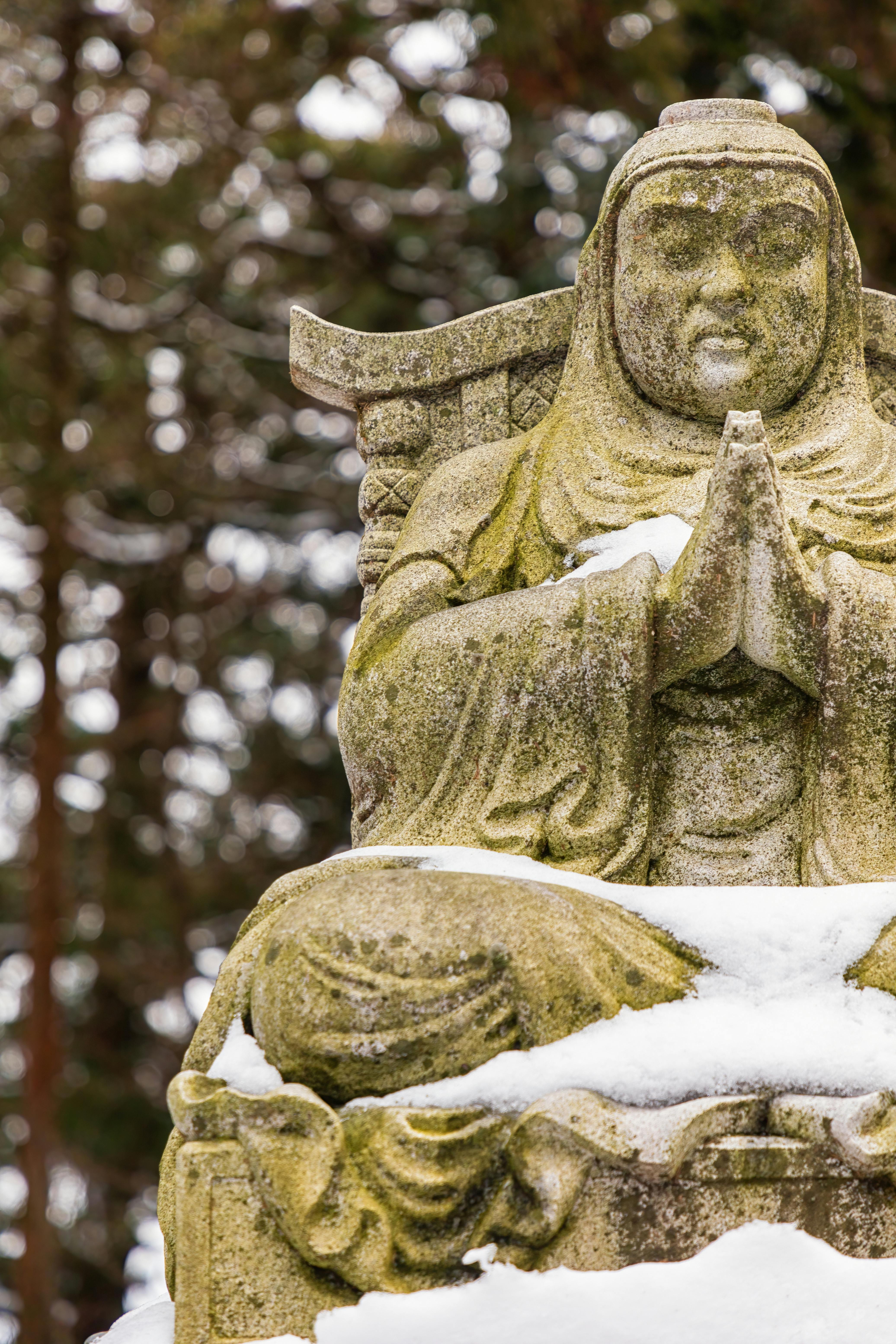 A stone statue of a seated figure with hands in a prayer position, partially covered in snow, set outdoors with blurred trees in the background.