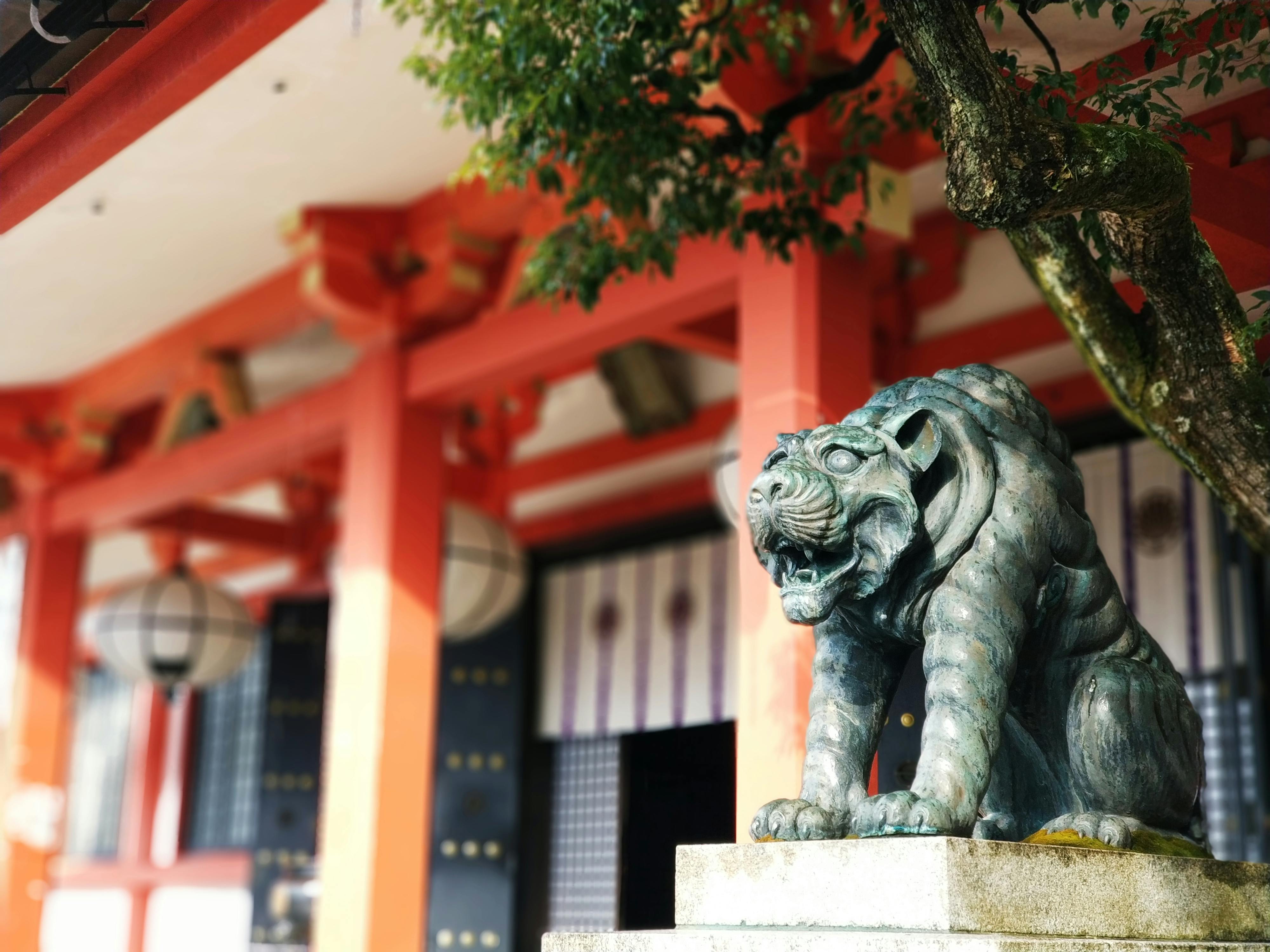 A bronze tiger statue stands on a stone pedestal in front of a traditional Japanese building with red pillars and white lanterns, partially shaded by a leafy tree.