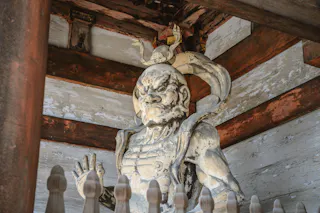 A weathered statue of a fierce guardian deity with a muscular build and an intense expression stands behind a wooden fence under a worn, wooden roof.