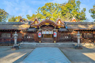 A traditional Japanese Shinto shrine with wooden architecture, ornate curved roof, stone lanterns, and lanterns decorated with red emblems, surrounded by trees under a blue sky.