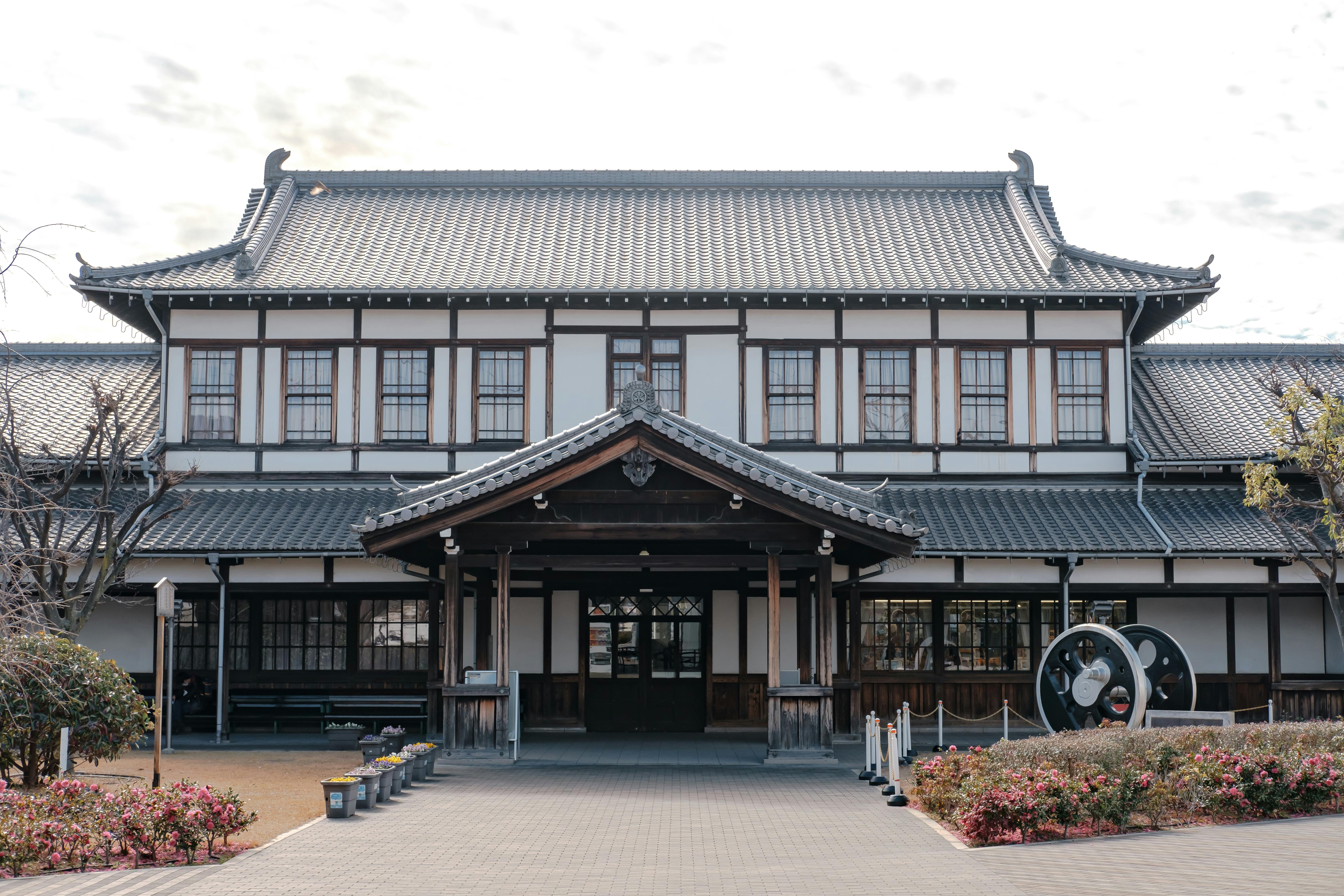 A traditional Japanese building with a tiled roof, wooden beams, and symmetrical windows, surrounded by bushes and a paved walkway leading to the entrance. There is a display with large wheels on the right side.