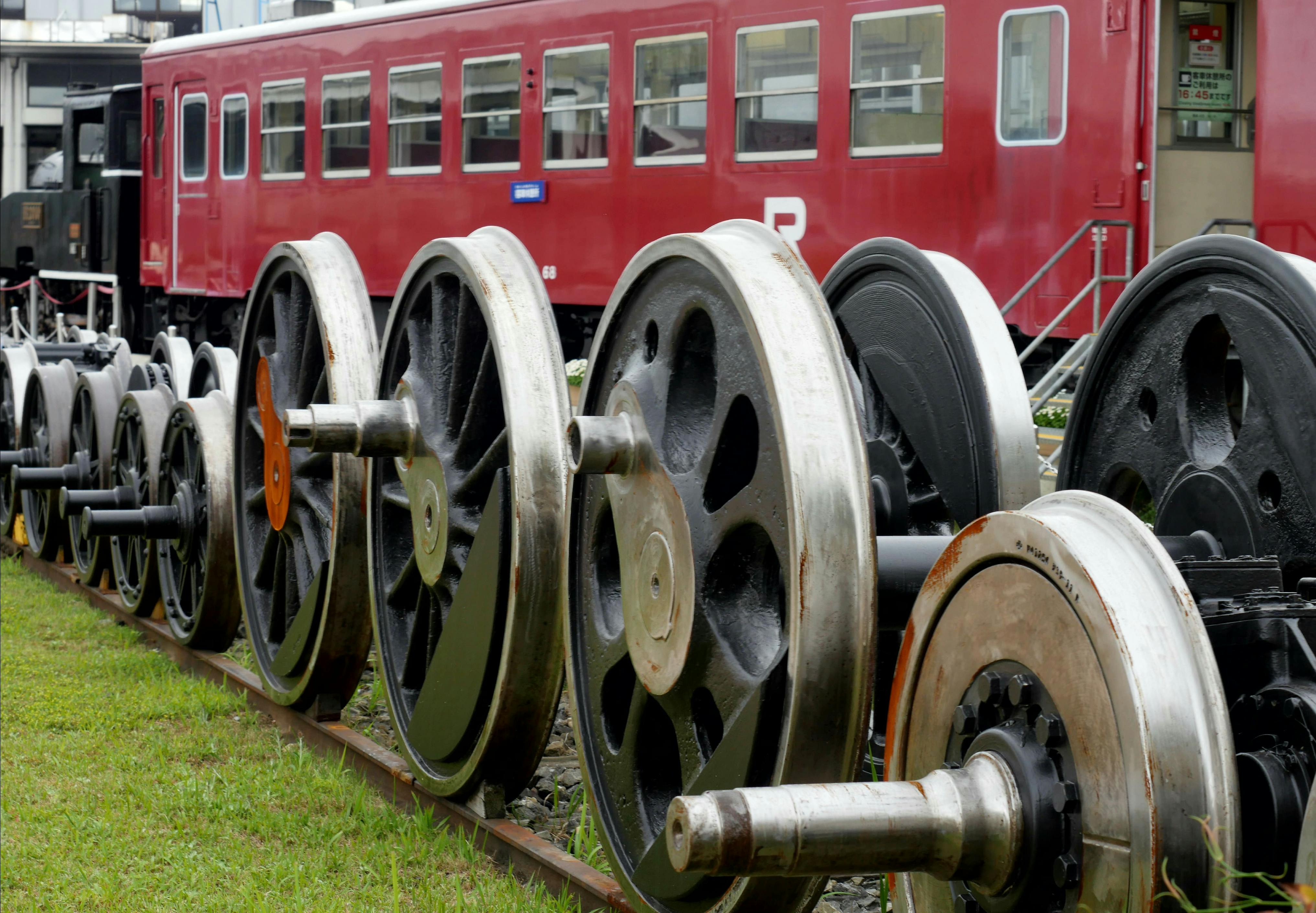 Several large, metal train wheels are lined up on a track in front of a stationary red passenger train car. Grass and a building are visible in the background.