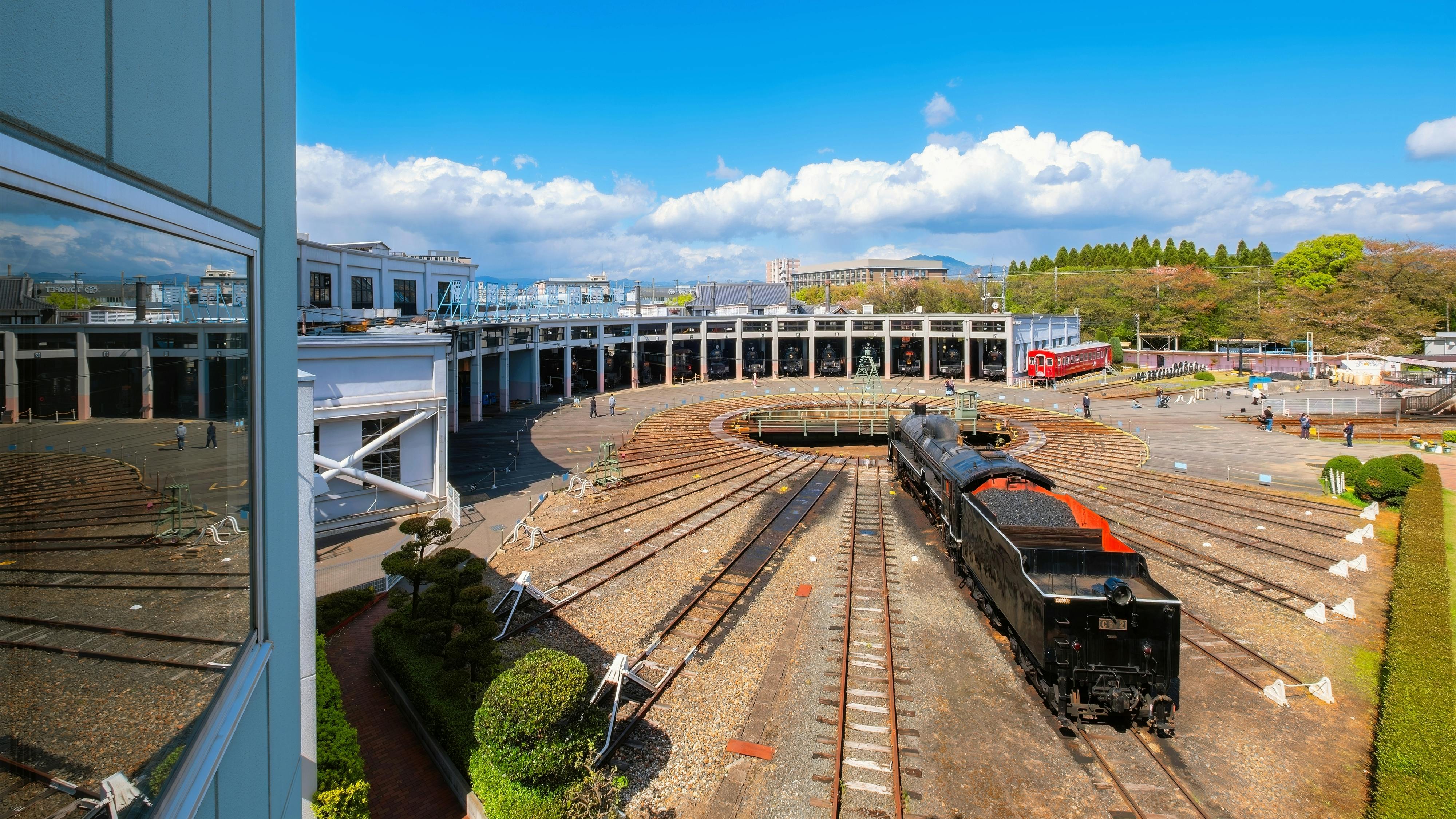 A black steam locomotive sits on a railway turntable surrounded by multiple train tracks, in front of a large roundhouse building under a bright blue sky with some clouds.
