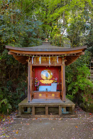 A small wooden Shinto shrine with a peaked roof stands in a lush, green forest. The shrine is decorated with flowers, white paper streamers, and an emblem, with stone steps visible to the right.